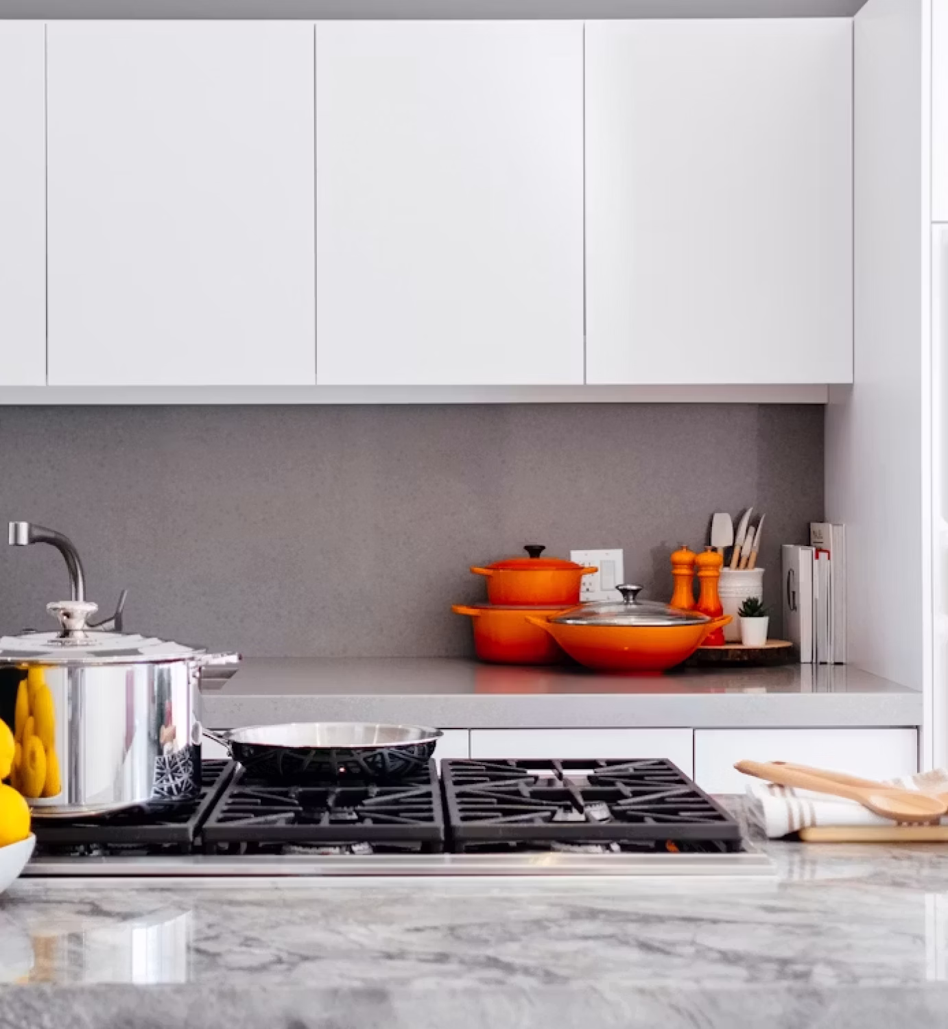 Modern kitchen with white cabinets, grey backsplash, stove with pots, and orange cookware on counter.
