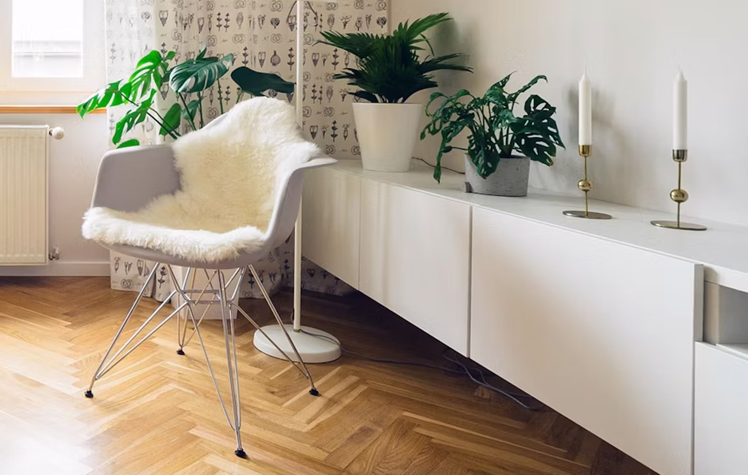 Modern grey chair with white fur throw next to plants and white sideboard in a bright room.