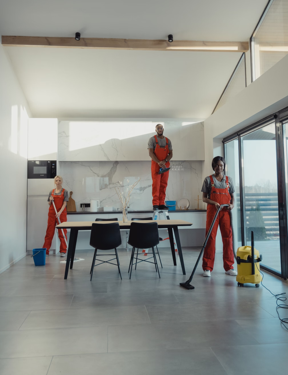 Three janitors in red overalls cleaning a modern kitchen with mop, vacuum, and window squeegee.