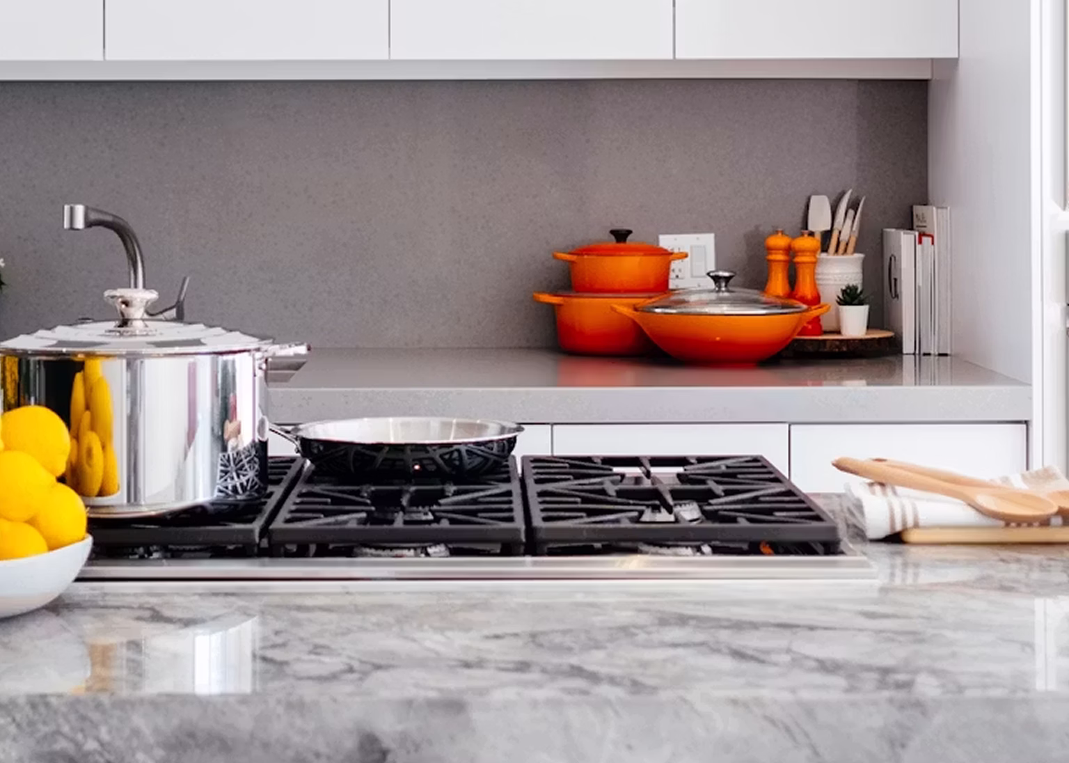 Modern kitchen stovetop with stainless steel pots, orange cookware, lemons, and wooden utensils.