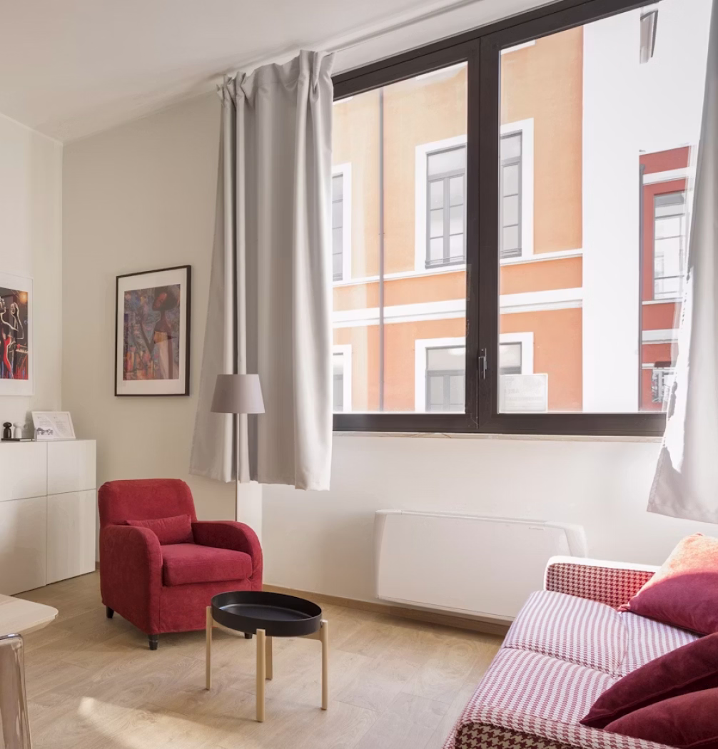Bright room with red armchair, patterned sofa, round black tray table, and large window with white curtains.