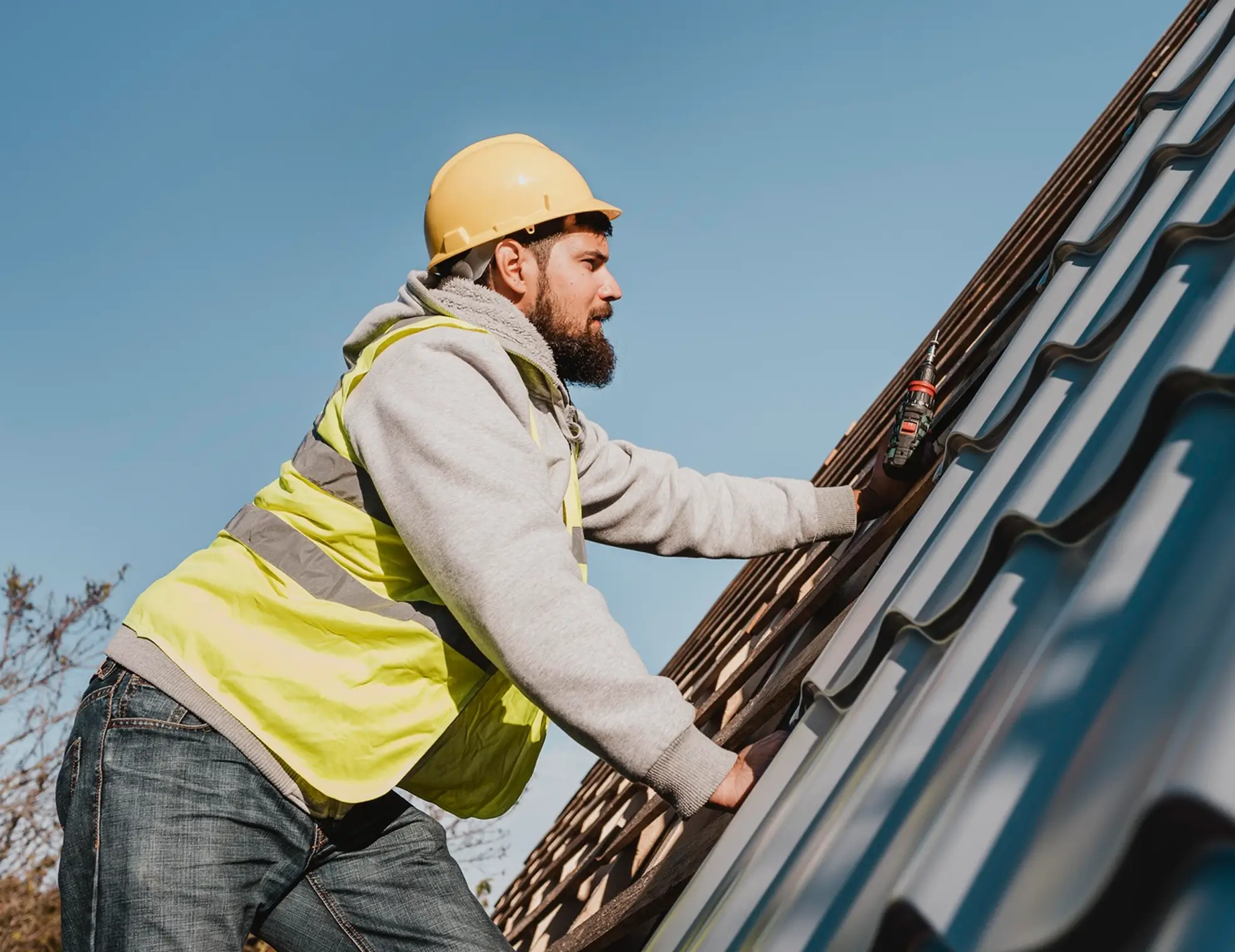 Construction worker in a yellow vest and helmet fixing metal roofing with a drill under clear sky.