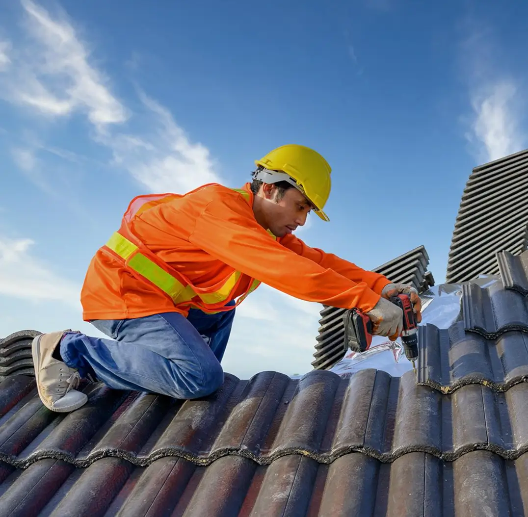 Construction worker in orange jacket and yellow helmet fixing roof tiles with a drill under blue sky.
