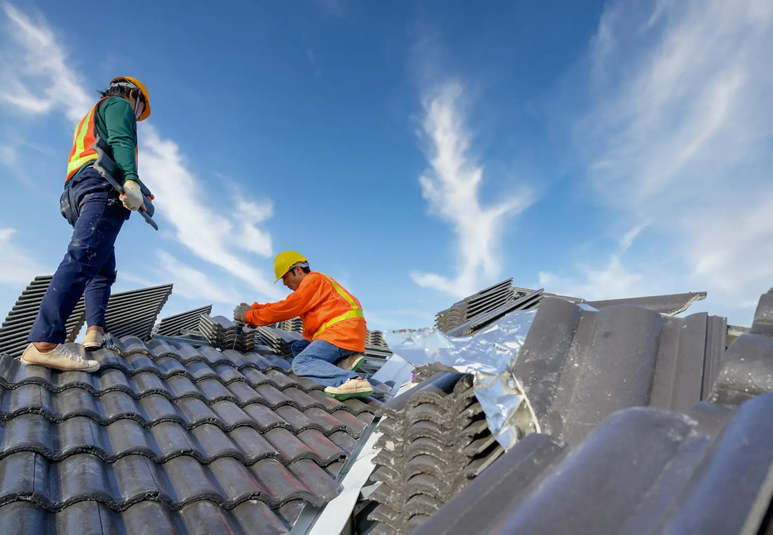 Two construction workers wearing helmets and safety vests installing roof tiles under clear sky.