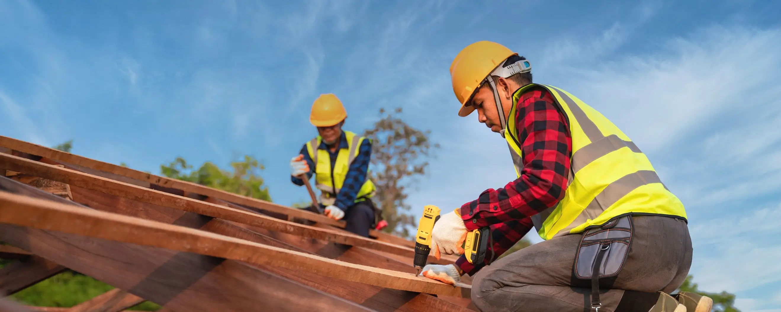 Two construction workers in safety gear working with wood on a roof frame under a blue sky.