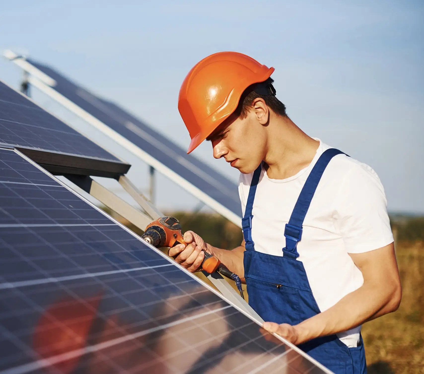 Worker wearing orange helmet and blue overalls installing solar panels with a drill outdoors.