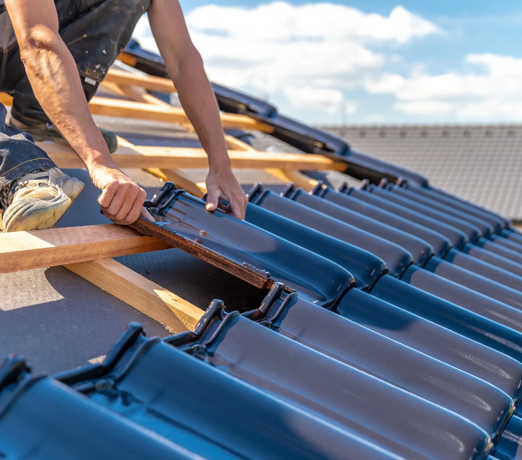 Worker installing blue ceramic roof tiles on wooden beams under a blue sky with clouds.