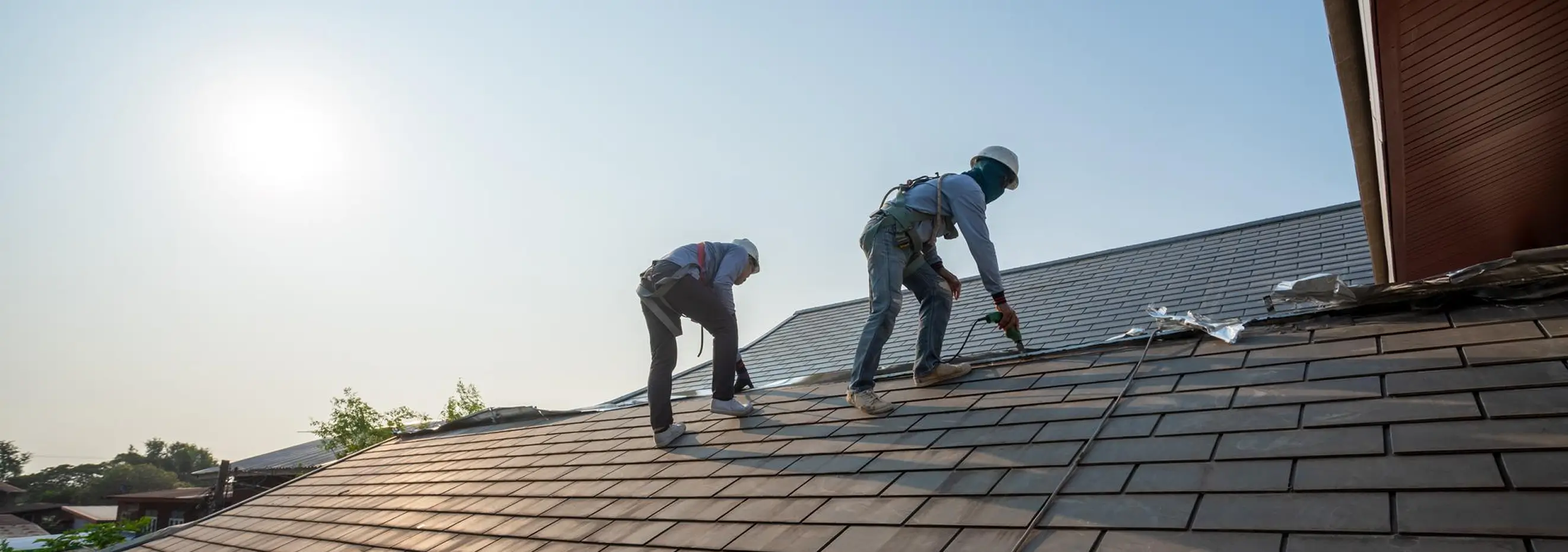 Two construction workers wearing helmets and safety harnesses work on a tiled roof under the sun.