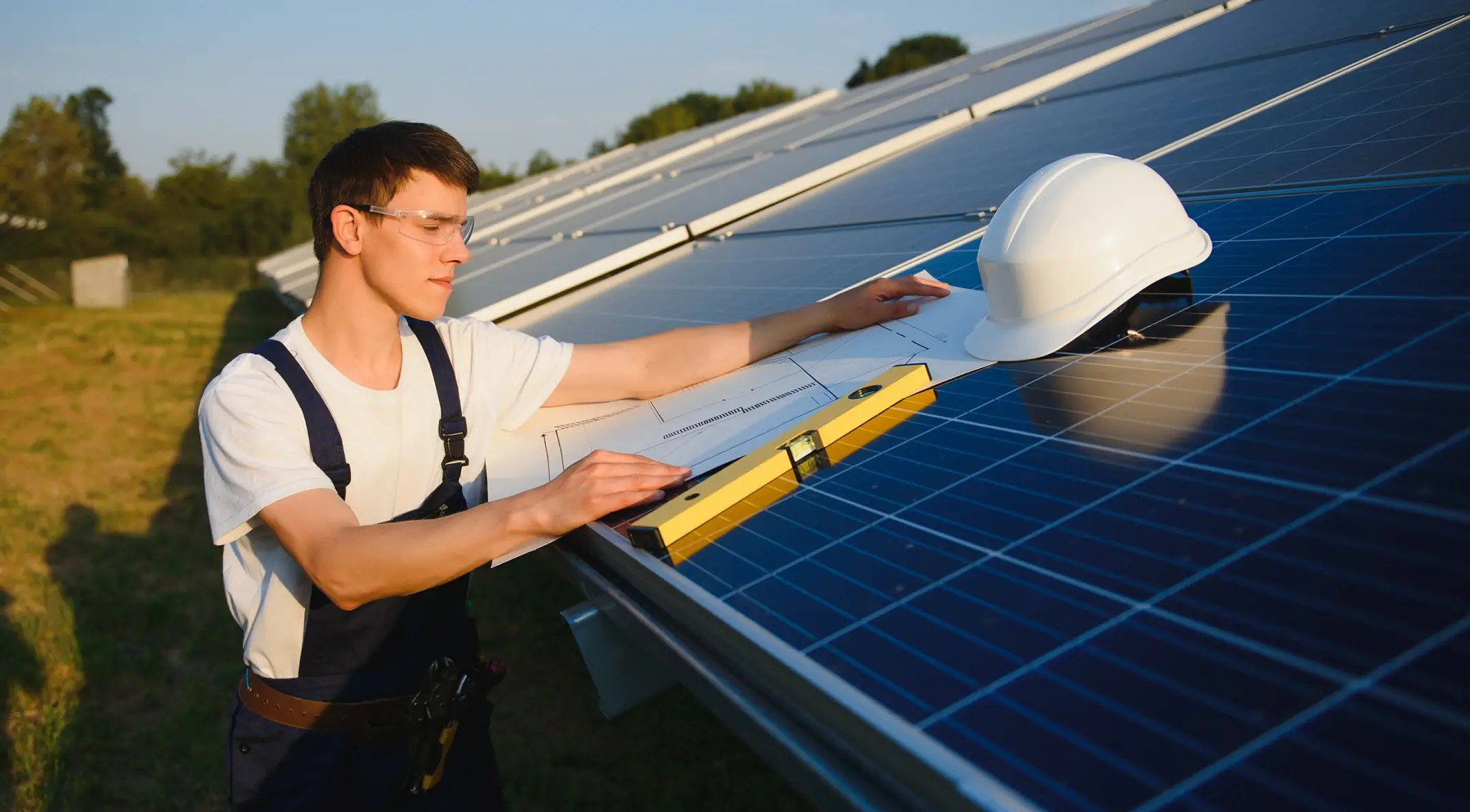 Engineer with safety glasses checking solar panels using a level and blueprint outdoors at sunset.