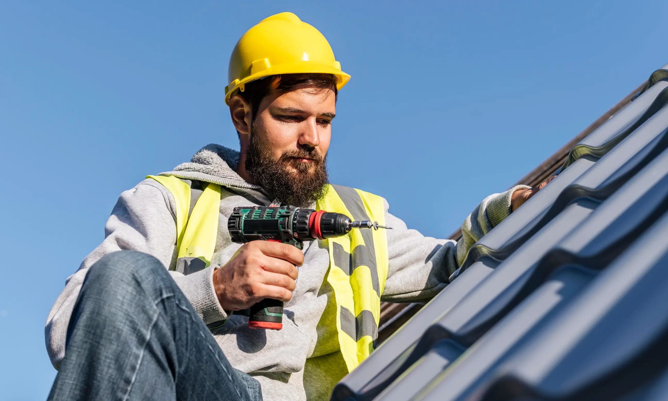 Bearded construction worker in a yellow hard hat and vest using a cordless drill on a metal roof.