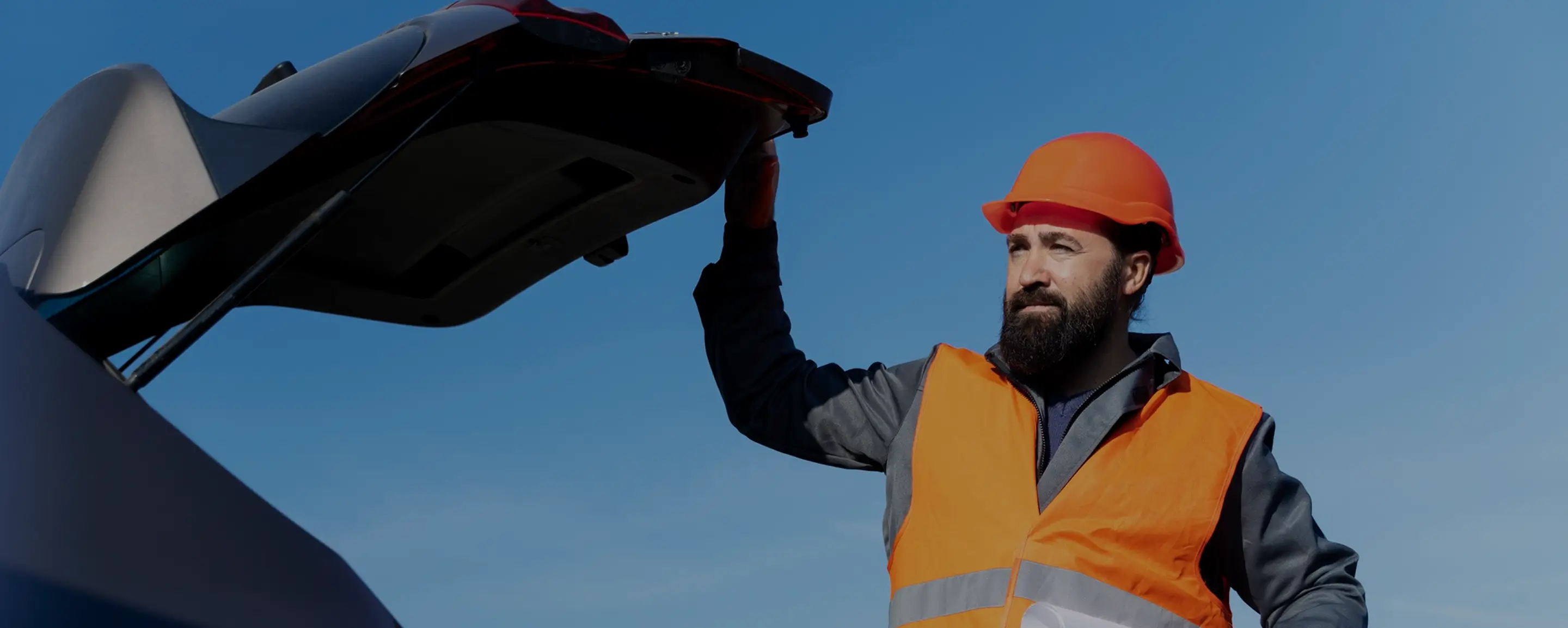 Man in orange safety vest and helmet opening a car trunk under a clear blue sky.