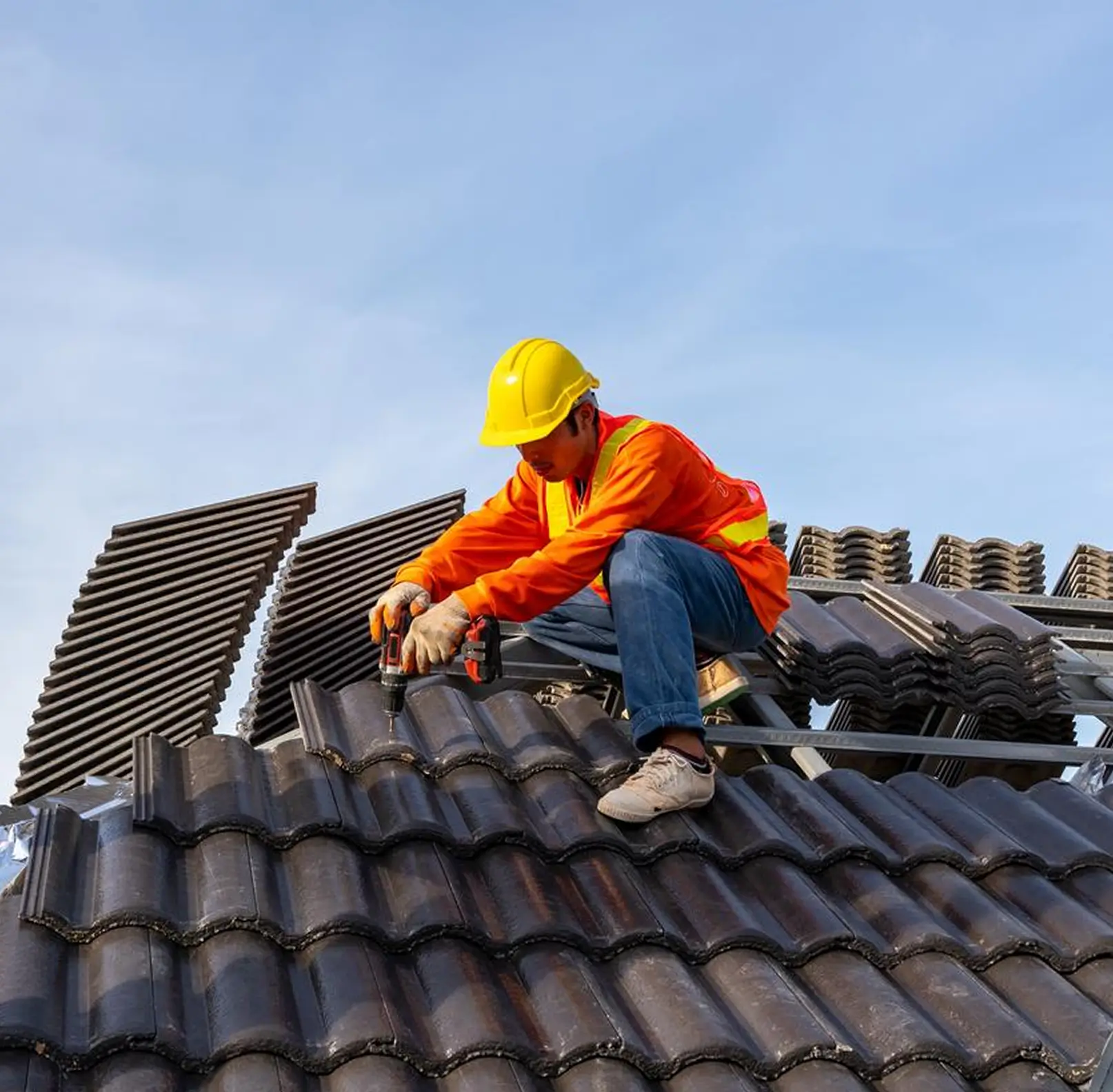 Construction worker in a yellow helmet and orange jacket installing dark roof tiles with a power drill.