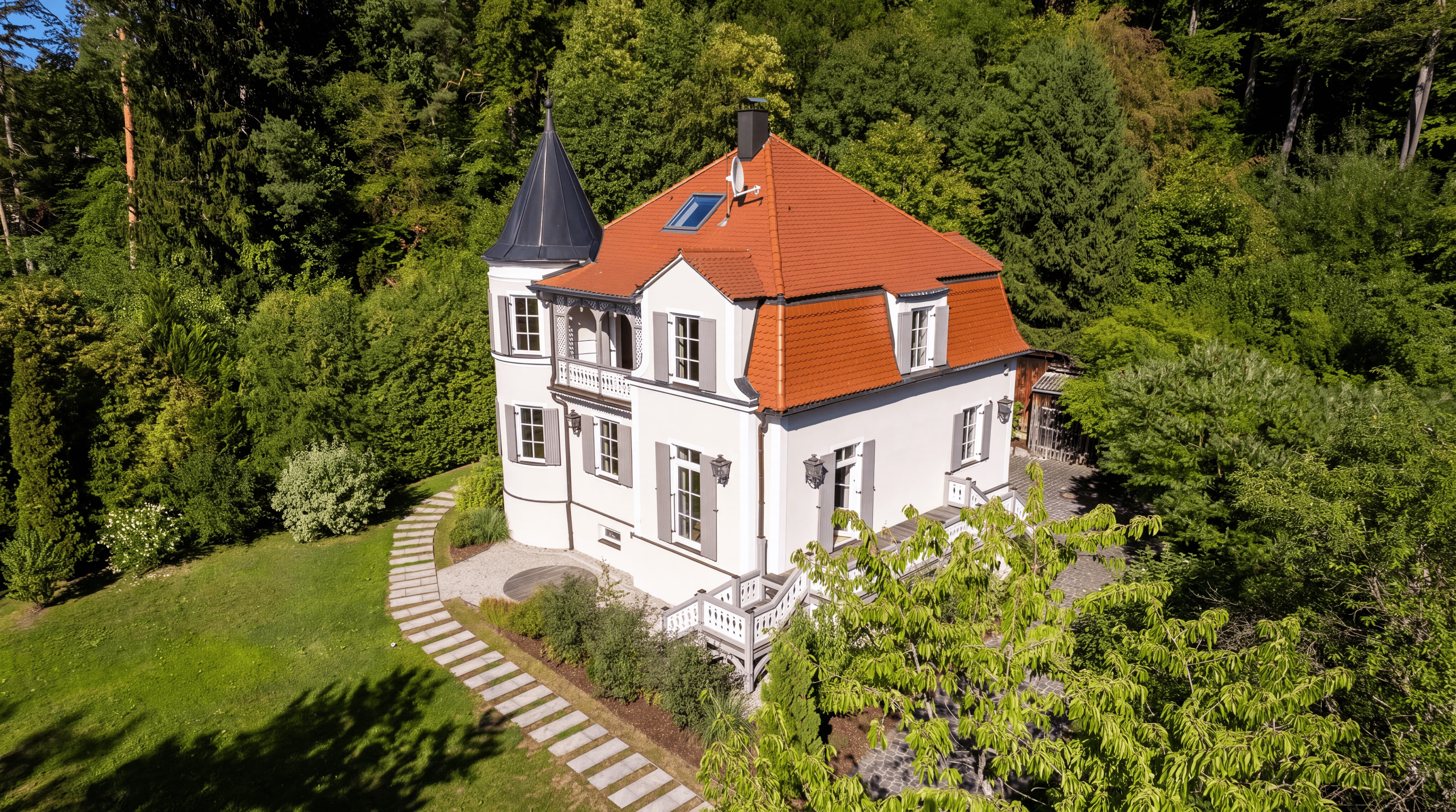 Weißes Haus mit rotem Ziegeldach und Turm, umgeben von grünem Wald und gepflegtem Garten.