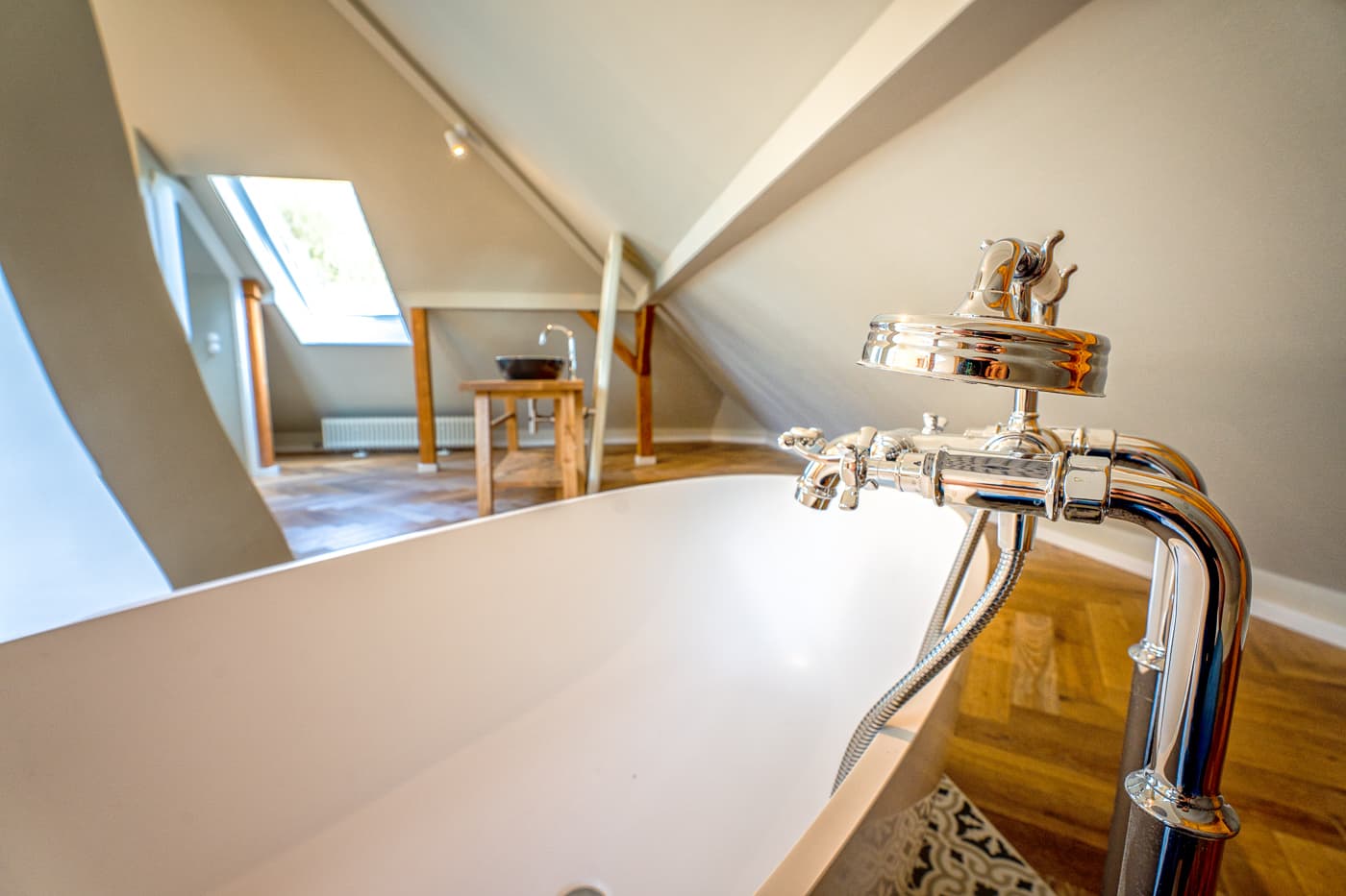 Freistehende Badewanne mit Chromarmatur in einem Dachzimmer mit Holzfußboden und Waschbecken.