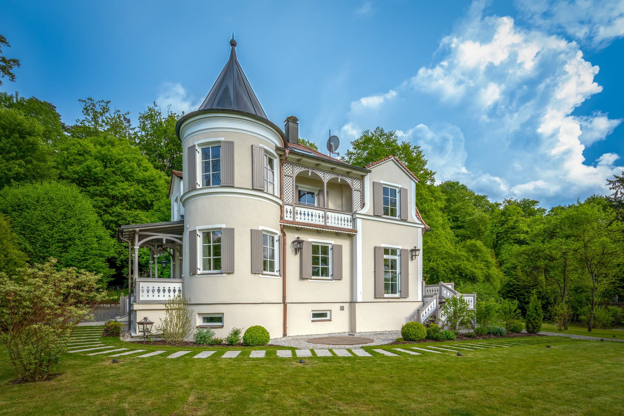 Elegantes Haus mit Turm und Balkon, umgeben von gepflegtem Garten und Bäumen unter blauem Himmel.