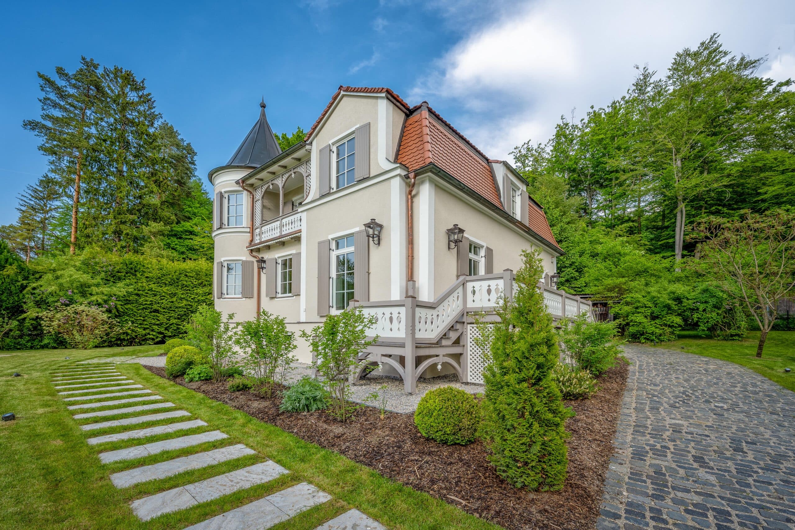 Elegantes Haus mit Turm, gepflegtem Garten, gepflastertem Weg und grünem Umfeld unter blauem Himmel.