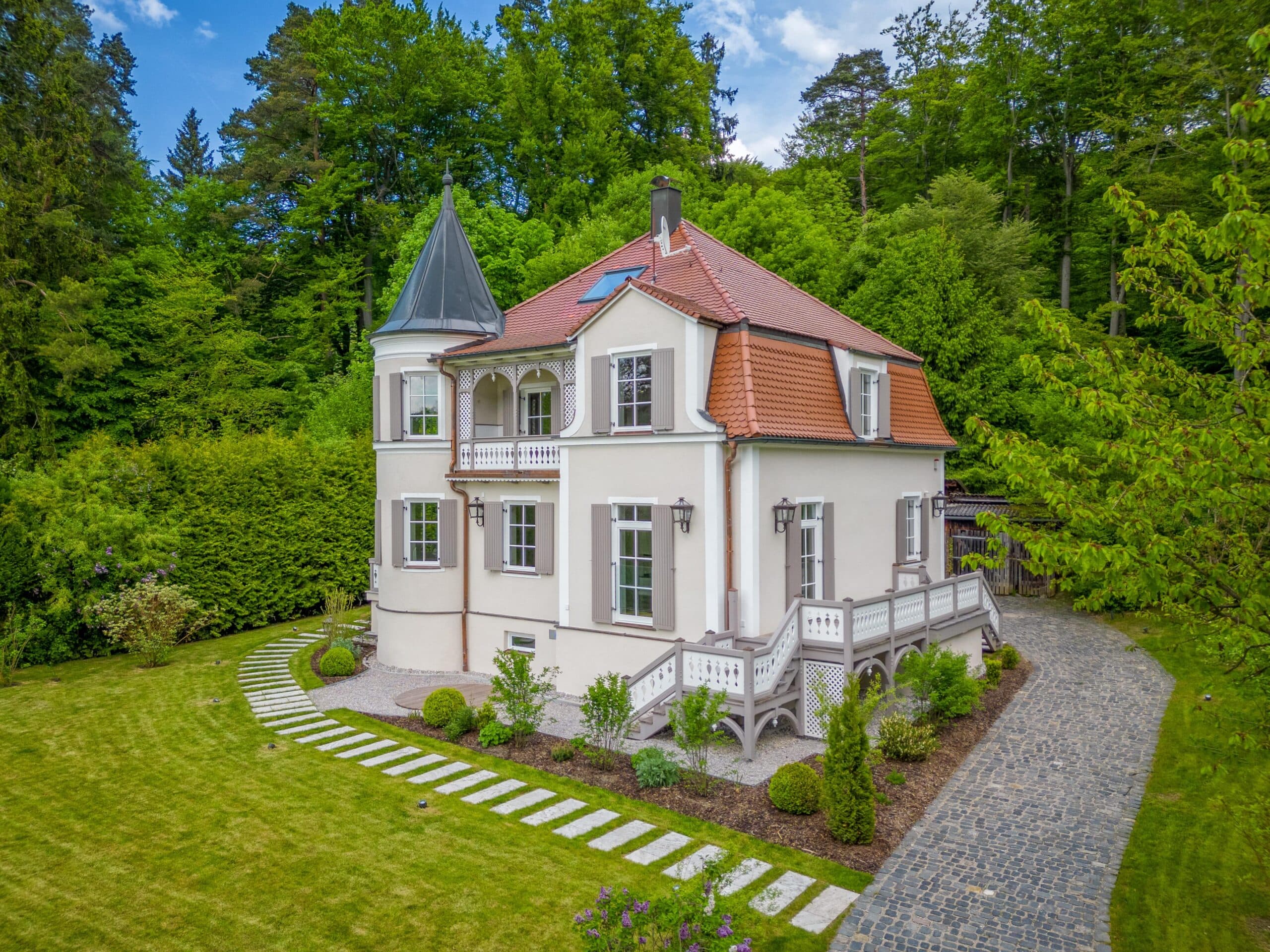Elegantes Haus mit Turm, rotem Ziegeldach und gepflegtem Garten am Waldrand.