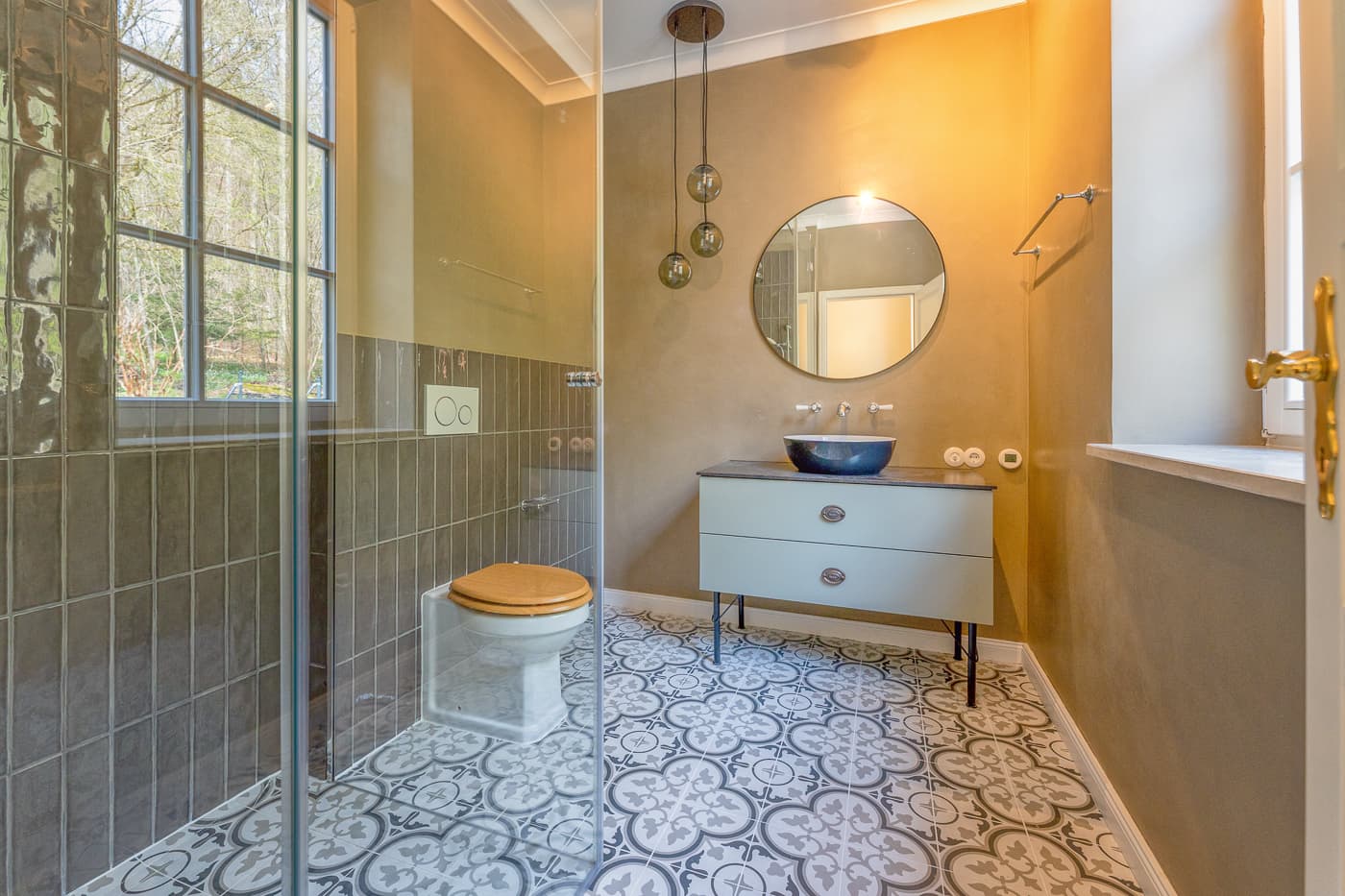 Modernes Badezimmer mit gemustertem Fliesenboden, grauer Toilette, schwarzem Waschbecken und rundem Spiegel.