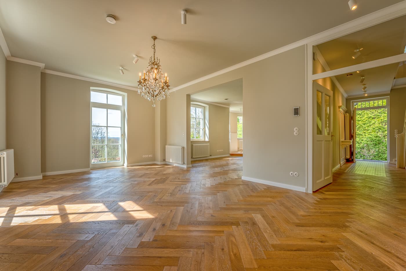 Helles, leeres Zimmer mit Parkettboden, Kronleuchter und großen Fenstern mit Gartenaussicht.