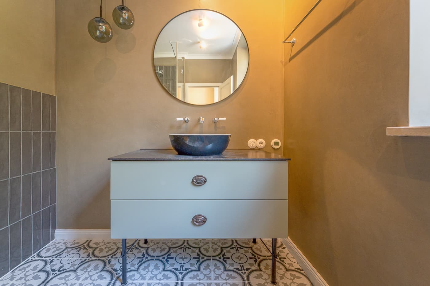 Modern bathroom sink with round mirror, hanging bulbs, and patterned floor tiles.