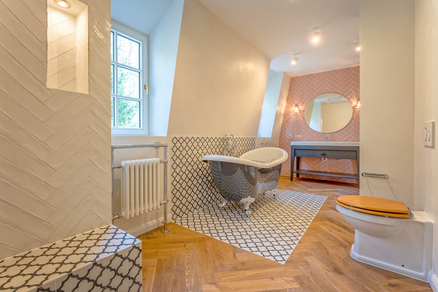 Modern bathroom with clawfoot bathtub on patterned tile and wooden floor, round mirror over sink.