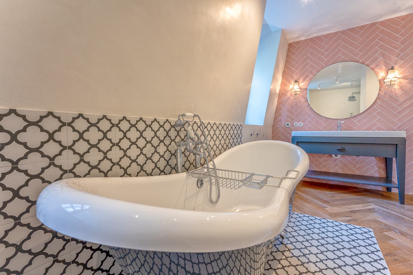 Freistehende Badewanne vor gemusterten Fliesen mit Spiegel und Waschbecken an rosa Wand.
