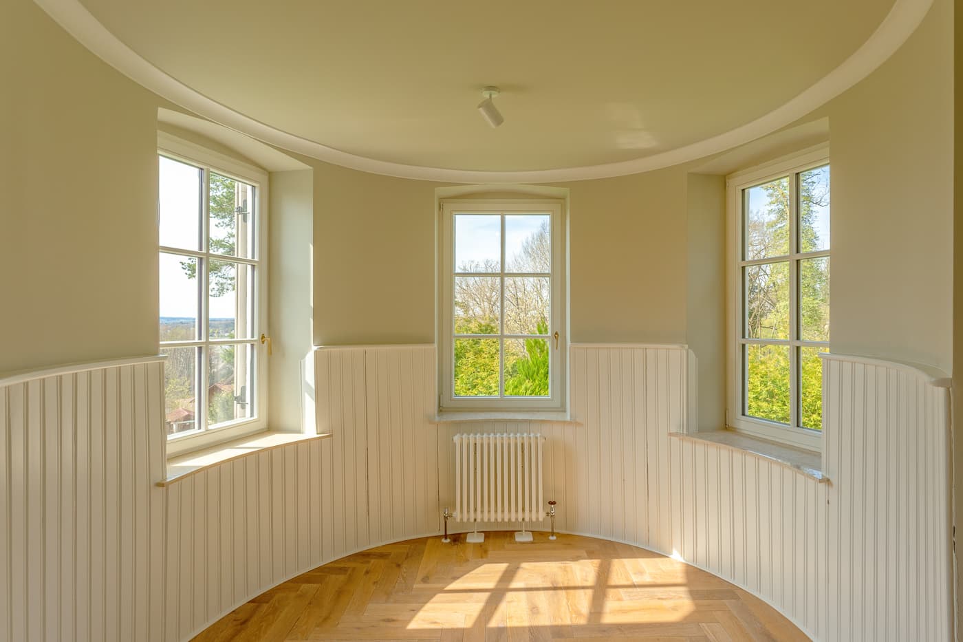 Runder Raum mit drei großen Fenstern, weißen Vertäfelungen und Parkettboden im Sonnenlicht.