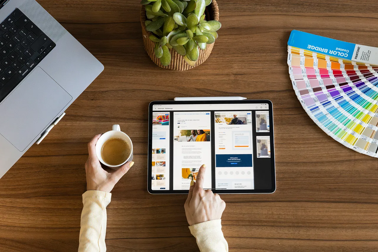 Een medewerker van Brandlos heeft een webdesign op staan op haar iPad. Ze wijst ernaar en heeft een kopje koffie vast. Verder ligt er een Pantone kleurenwaaier en een macbook