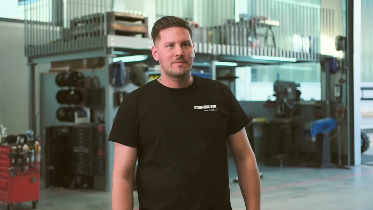 Simon steht in der Werkstatt und spricht in die Kamera