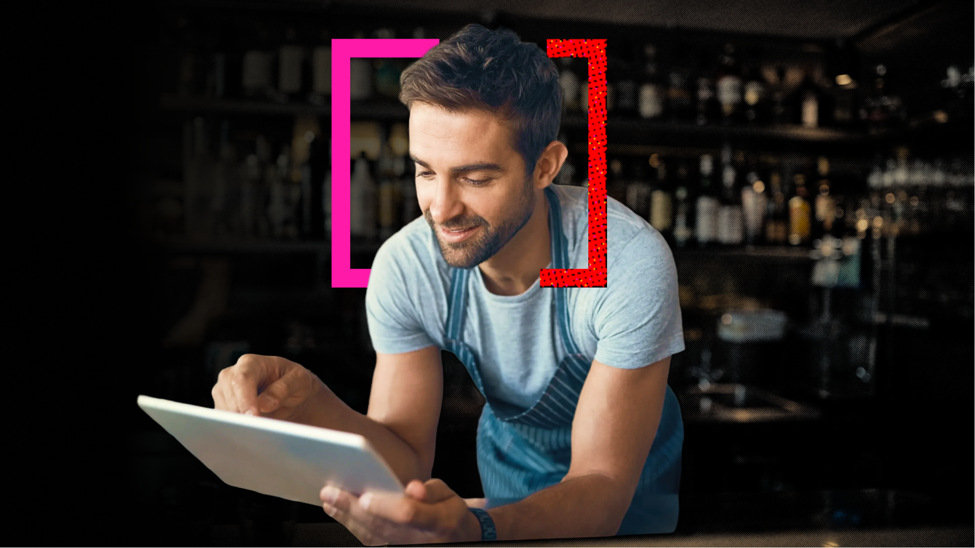 A man, leaning on a bar, wearing a grey shirt and blue apron, engaging and looking at his tablet.