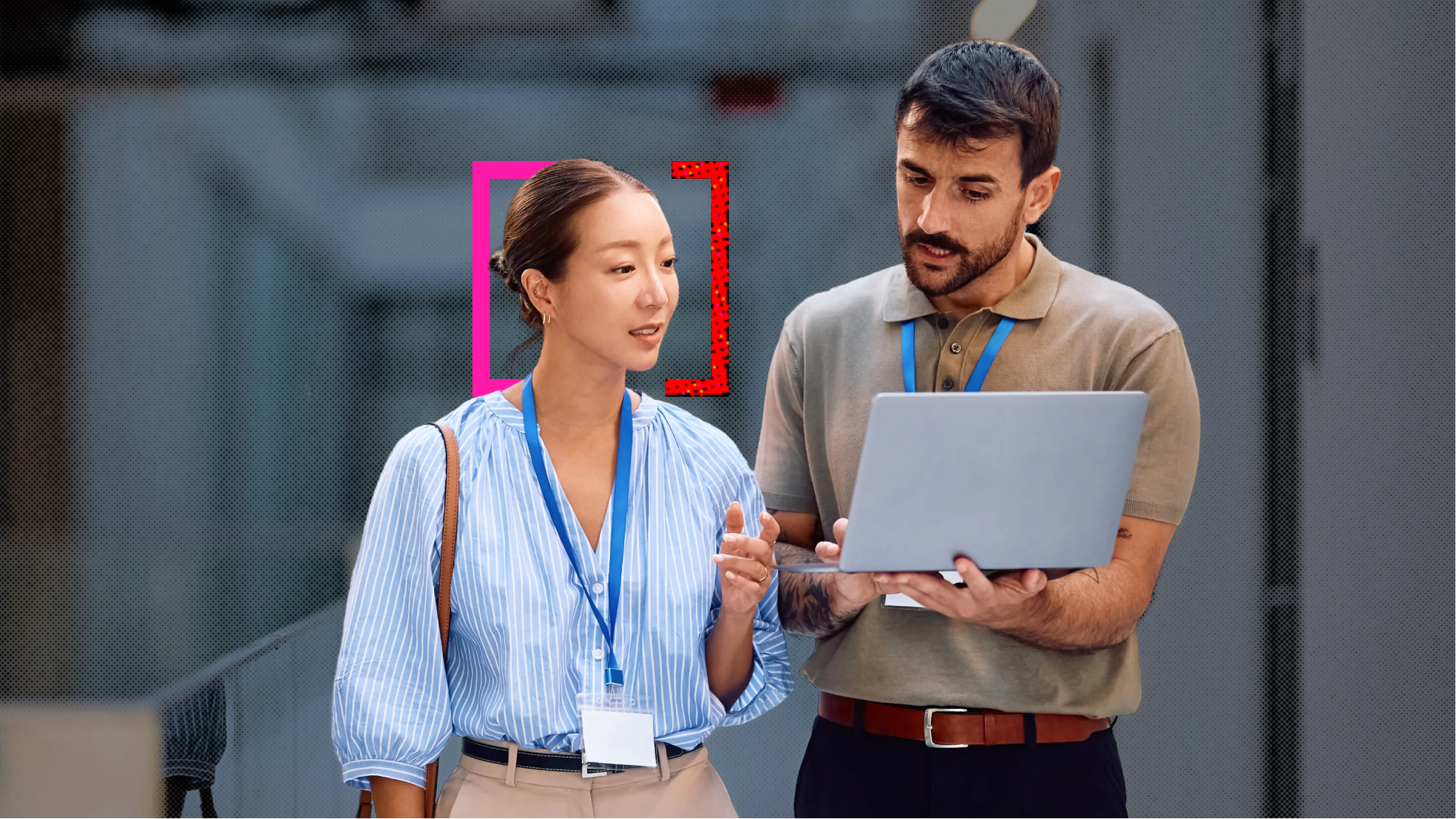 A man and a woman office workers, both wearing office attire and blue lanyards, standing and discussing something on the man's laptop.