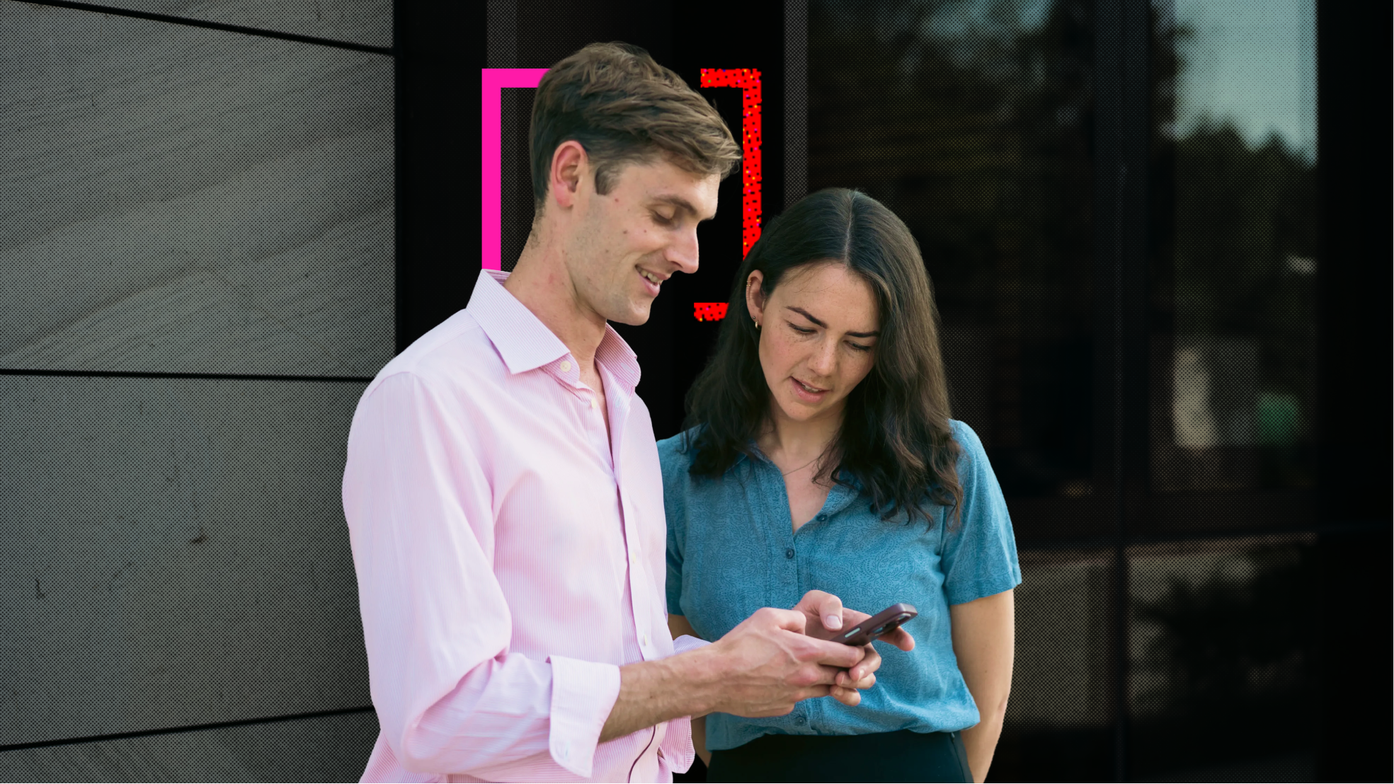 Two people, a man and a woman, outside a building in office attire, both looking at the man's phone in his hands.