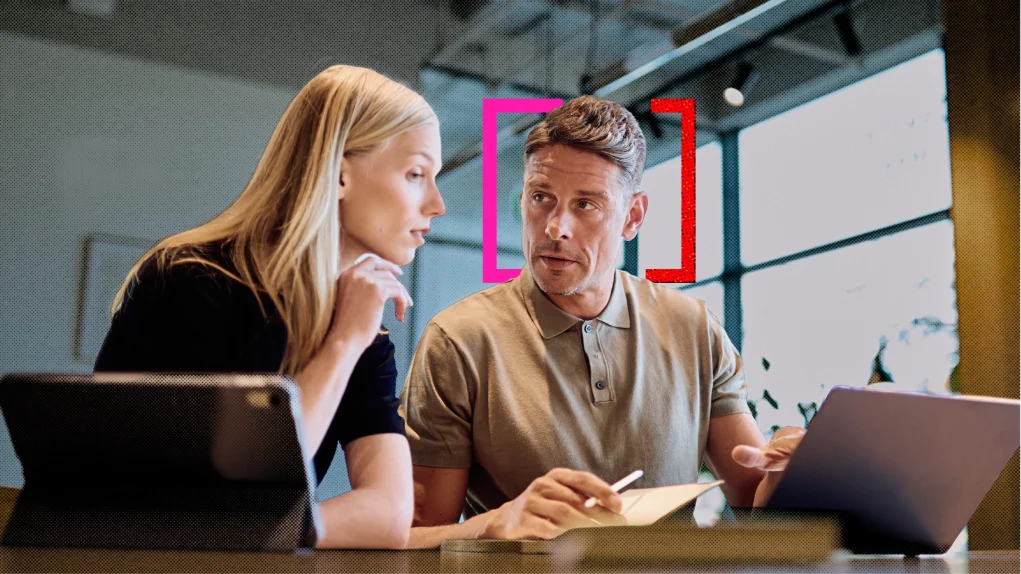 A woman and a man discuss notes beside a laptop in a bright modern office, leaning together with focused expressions; pink and red brackets frame him.