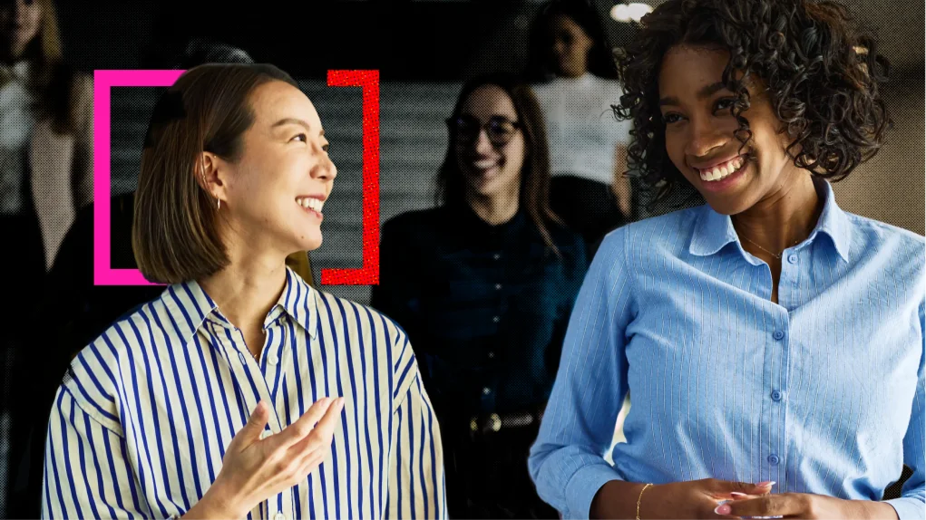 Two smiling women chat in a bright office, with colleagues in the background; pink and red corner brackets frame the woman on the left, conveying a warm, upbeat mood.