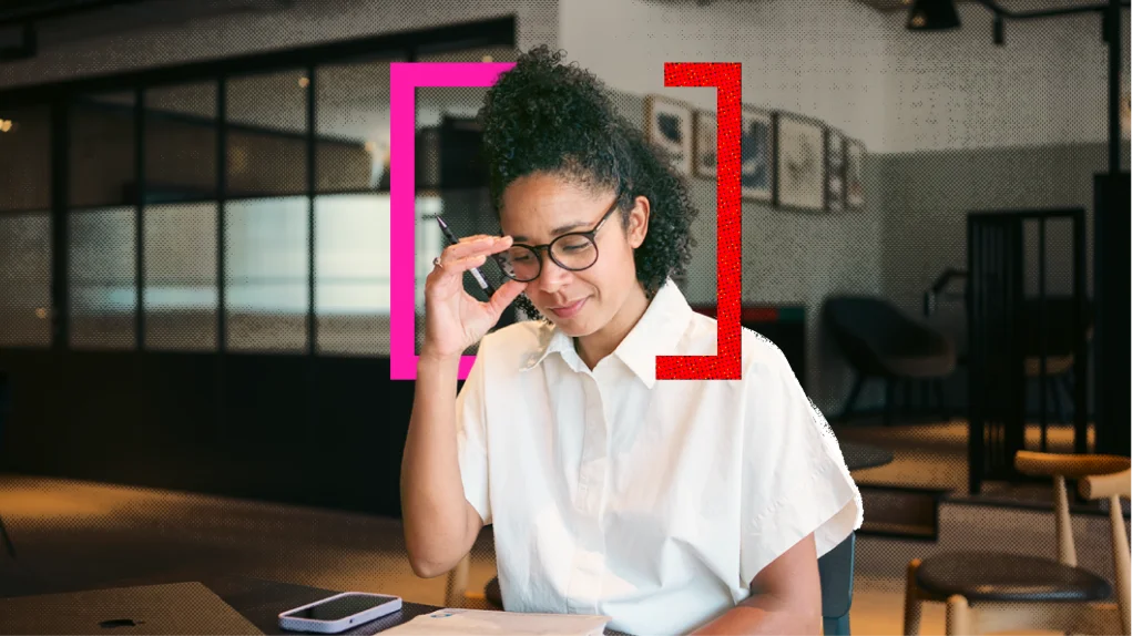 A woman in glasses adjusts them at an office desk, framed by bright pink and red corner brackets; she looks focused and thoughtful.