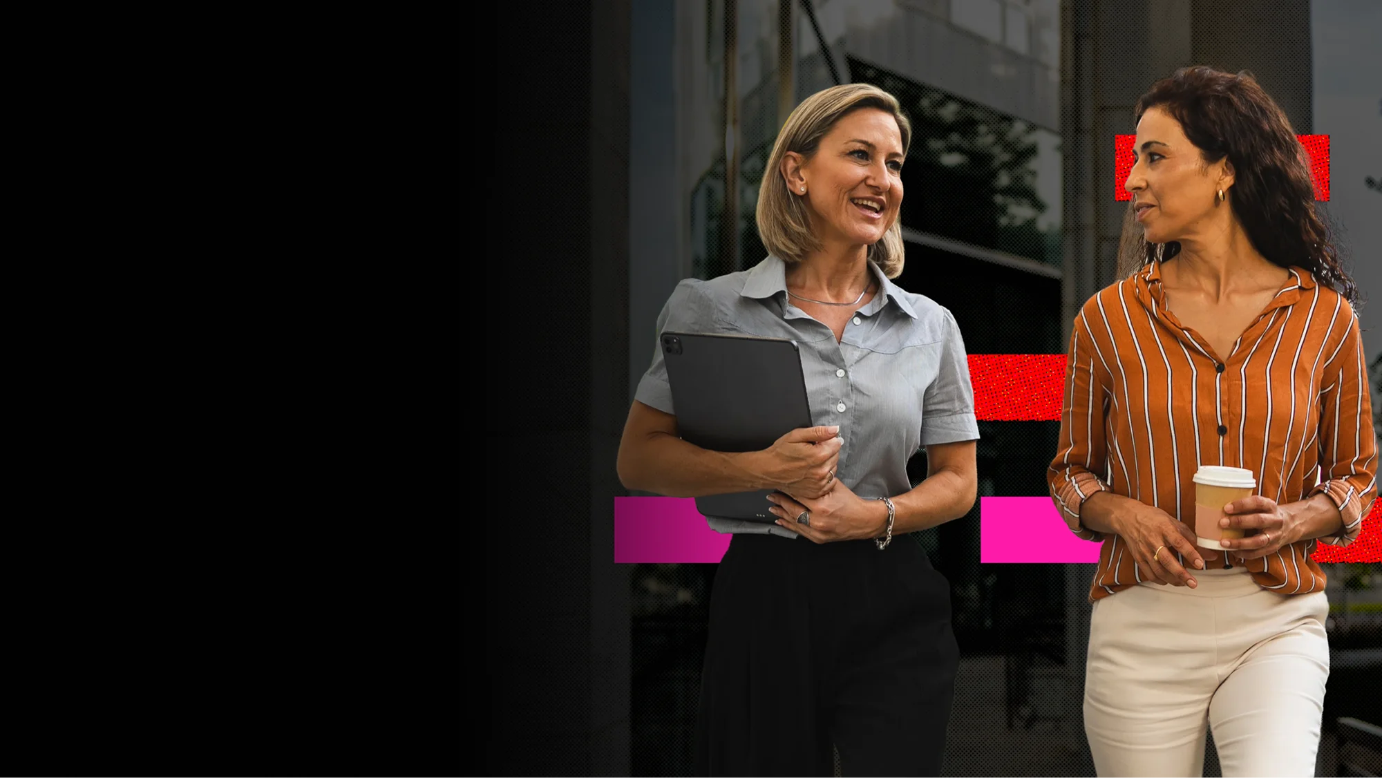 Two professional women walking and talking outdoors, one holding a tablet and the other holding a coffee cup.