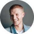 Smiling young man with short blond hair wearing a light denim jacket against a gray background.
