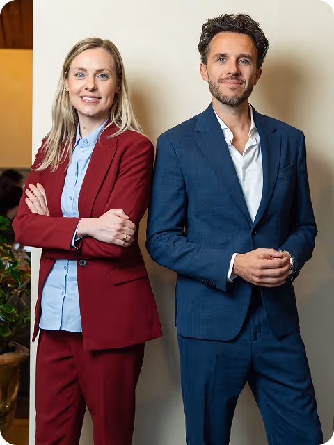 Two business professionals standing side by side, a woman in a red suit with arms crossed and a man in a blue suit with hands clasped.