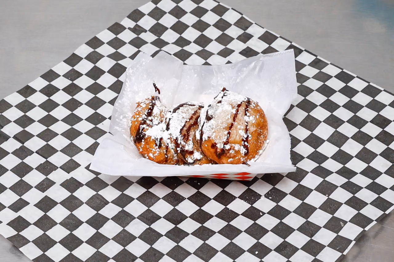 Three fried round desserts drizzled with chocolate and dusted with powdered sugar in a paper tray.