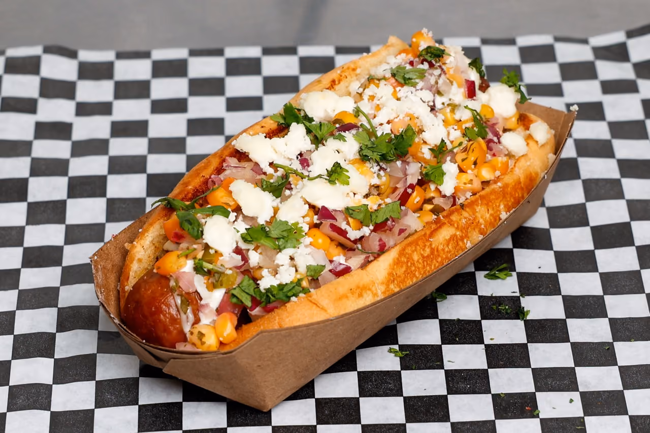 Hot dog topped with corn, diced onions, cilantro, white cheese, and creamy sauce in a paper tray.