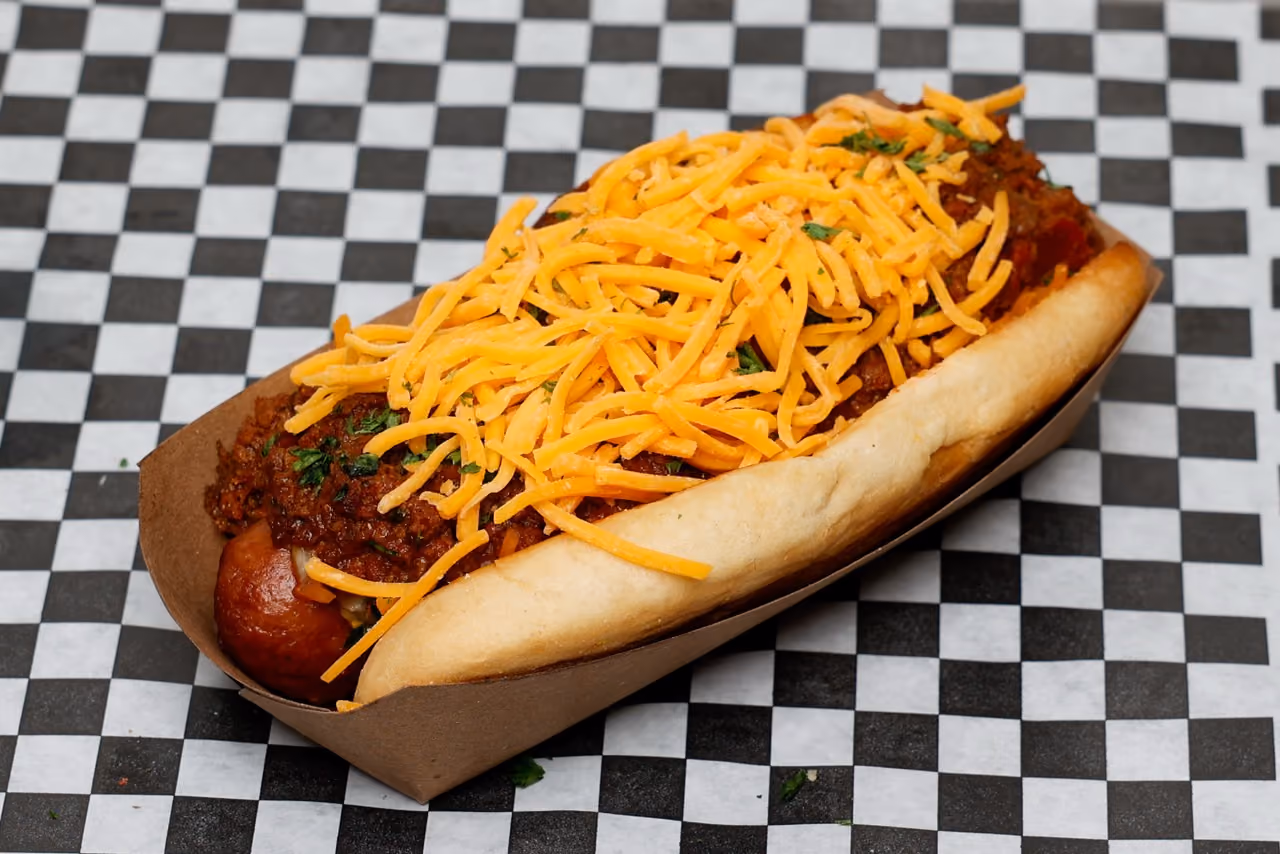 Chili cheese dog topped with shredded cheddar cheese on a black and white checkered paper.