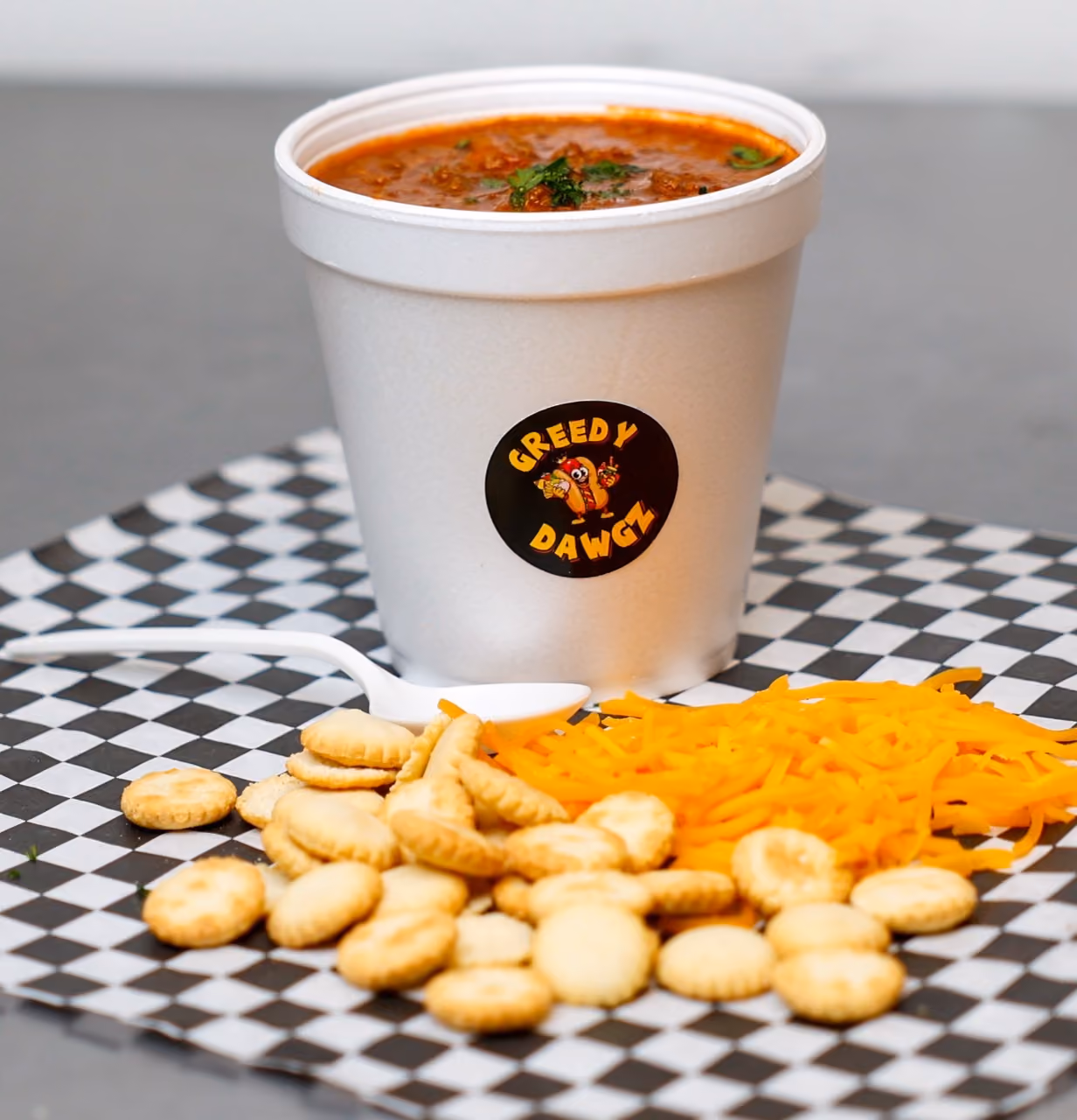 Cup of chili with crackers, shredded cheddar cheese, and a white plastic spoon on checkered paper.