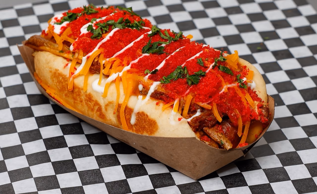 Hot dog with shredded cheddar, red crushed seasoning, chopped herbs, and white sauce in a paper tray.