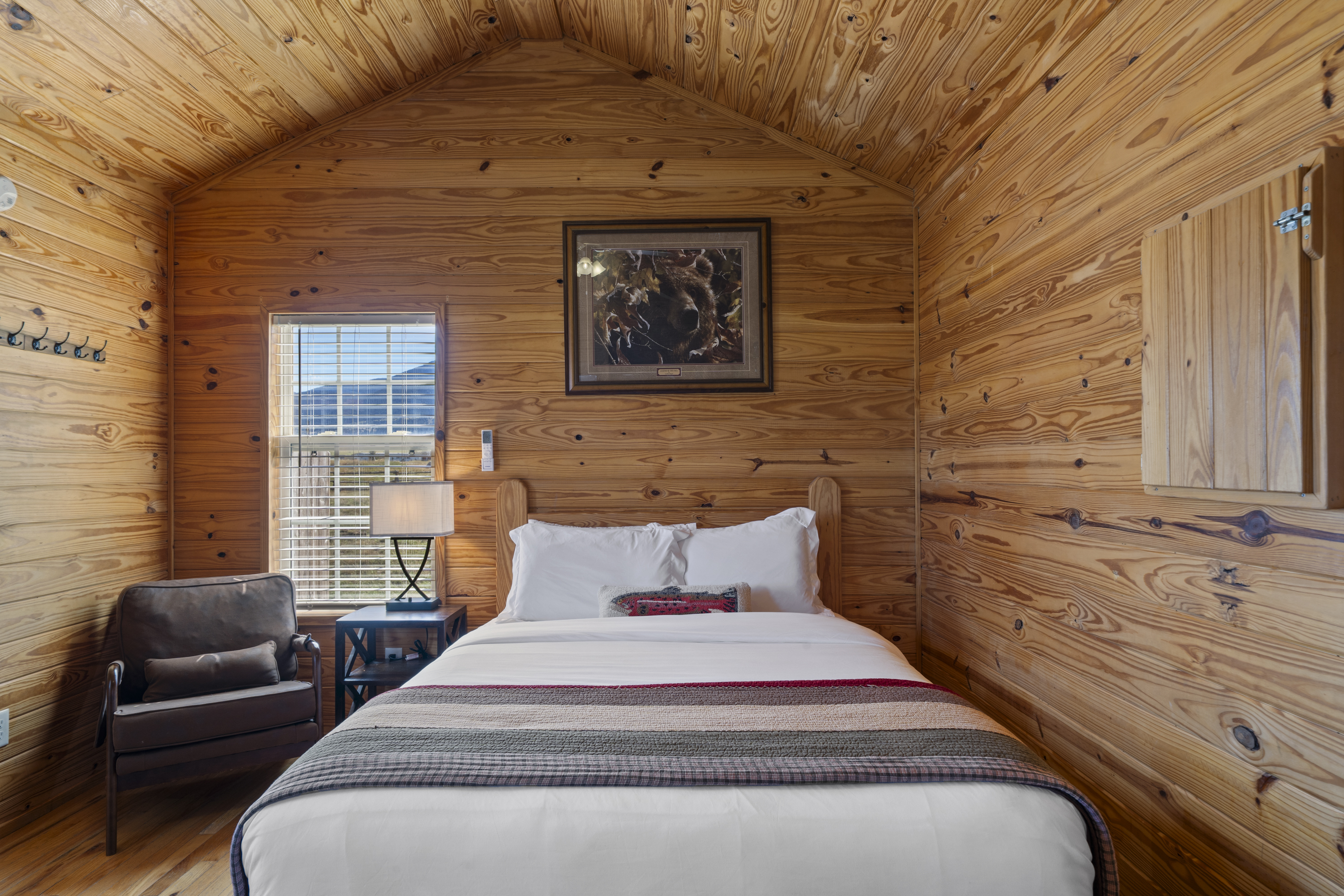 Cabin queen bed under a vaulted pine ceiling, with a reading chair, bedside lamp and bear print