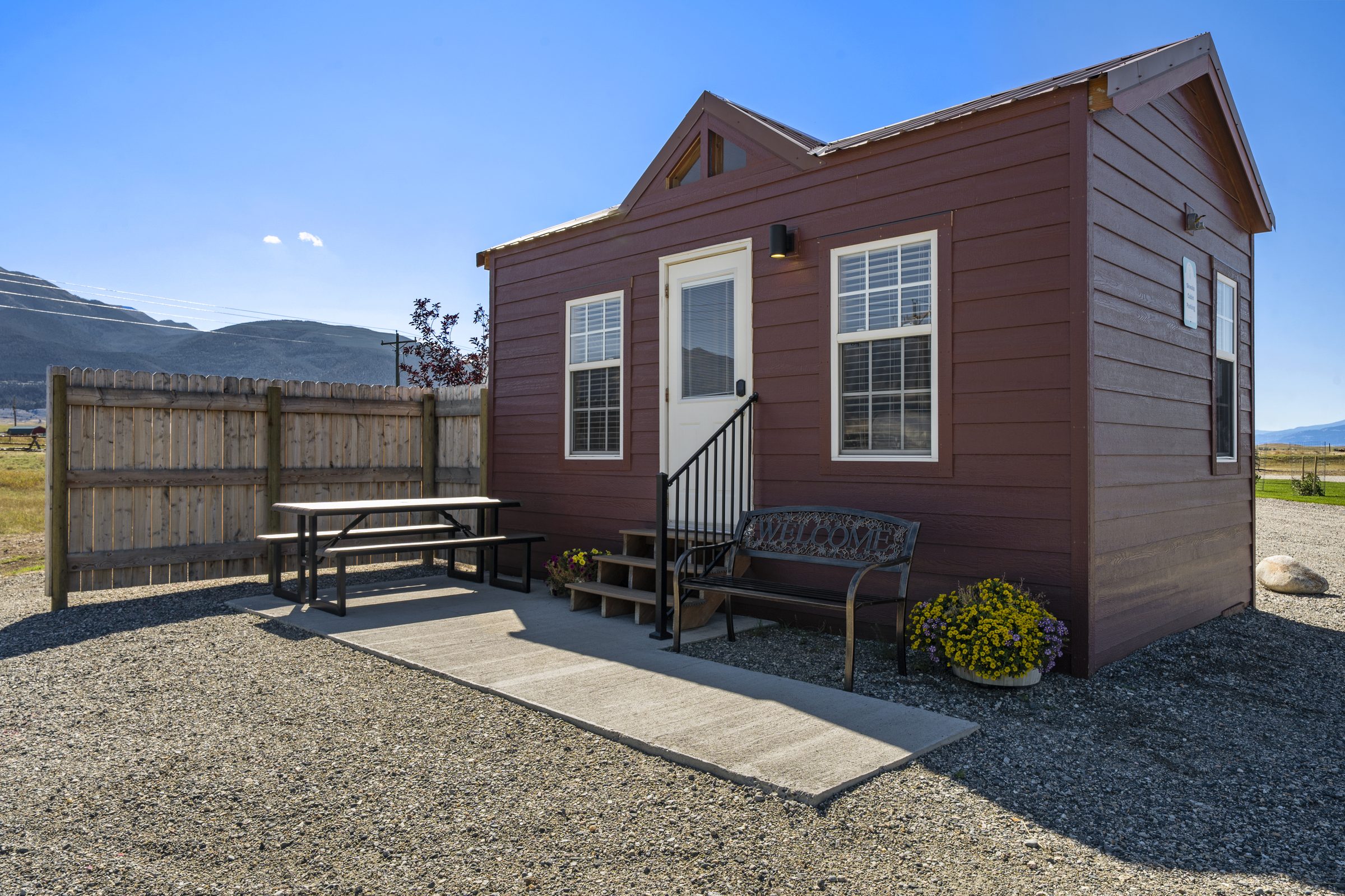 Skwala Cabin, with a concrete patio, picnic table and Welcome bench framed by flowers