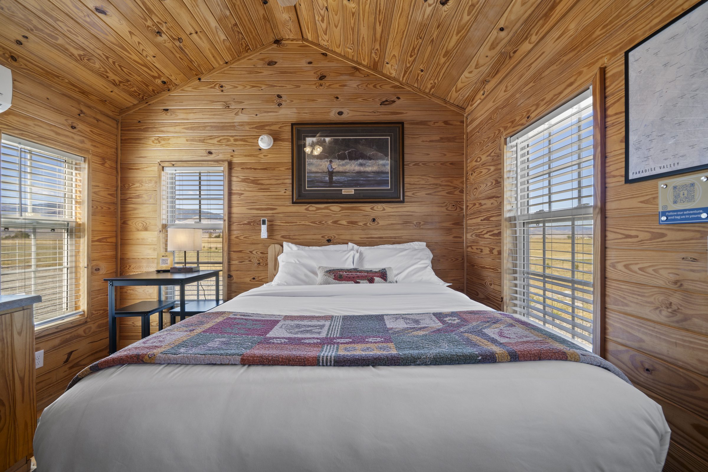 Queen bed under a vaulted pine ceiling, with fly-fishing art and windows on both sides