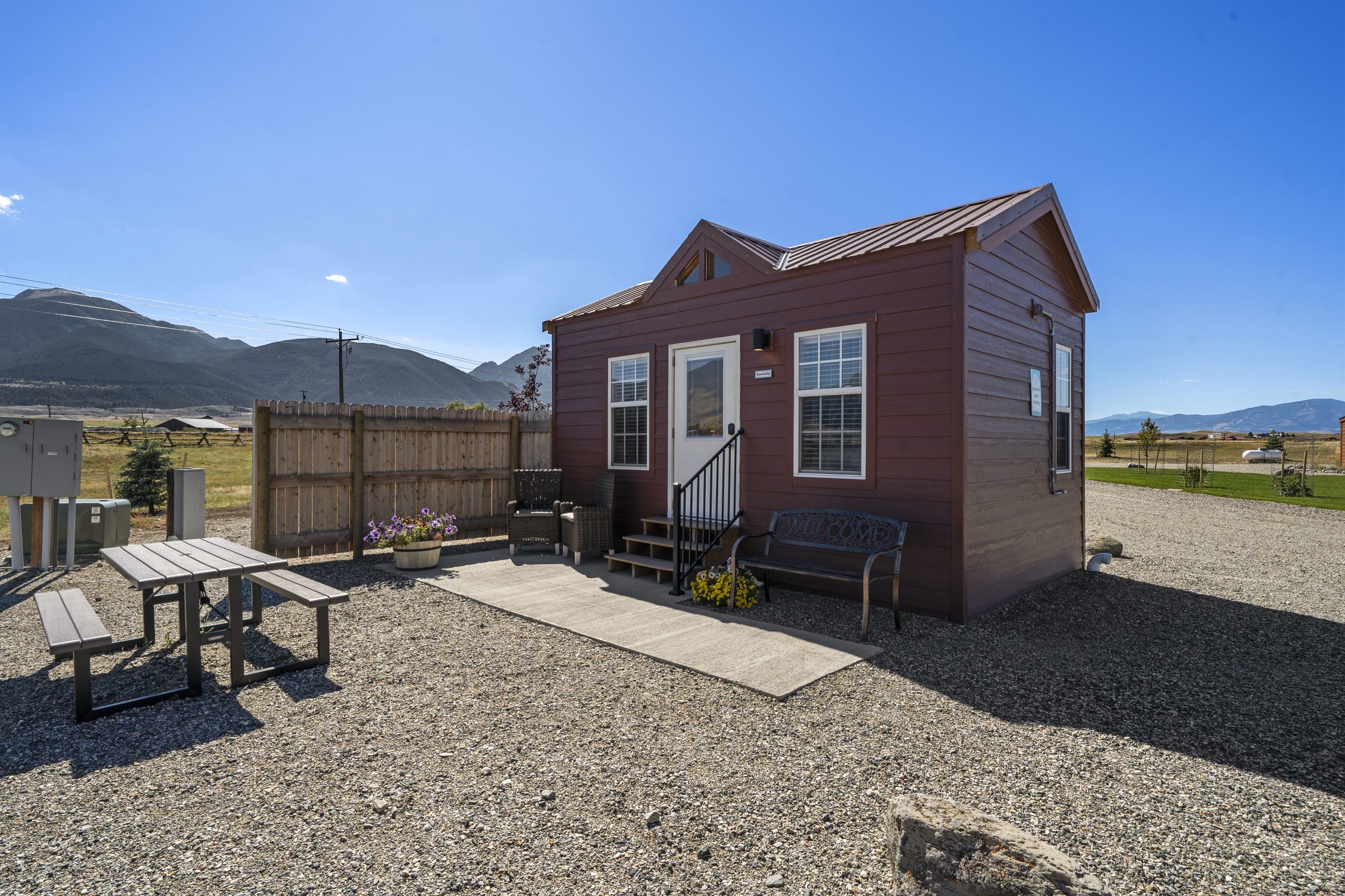 Damselfly Cabin, with a concrete patio, picnic table, two wicker chairs and a Welcome bench, mountains behind
