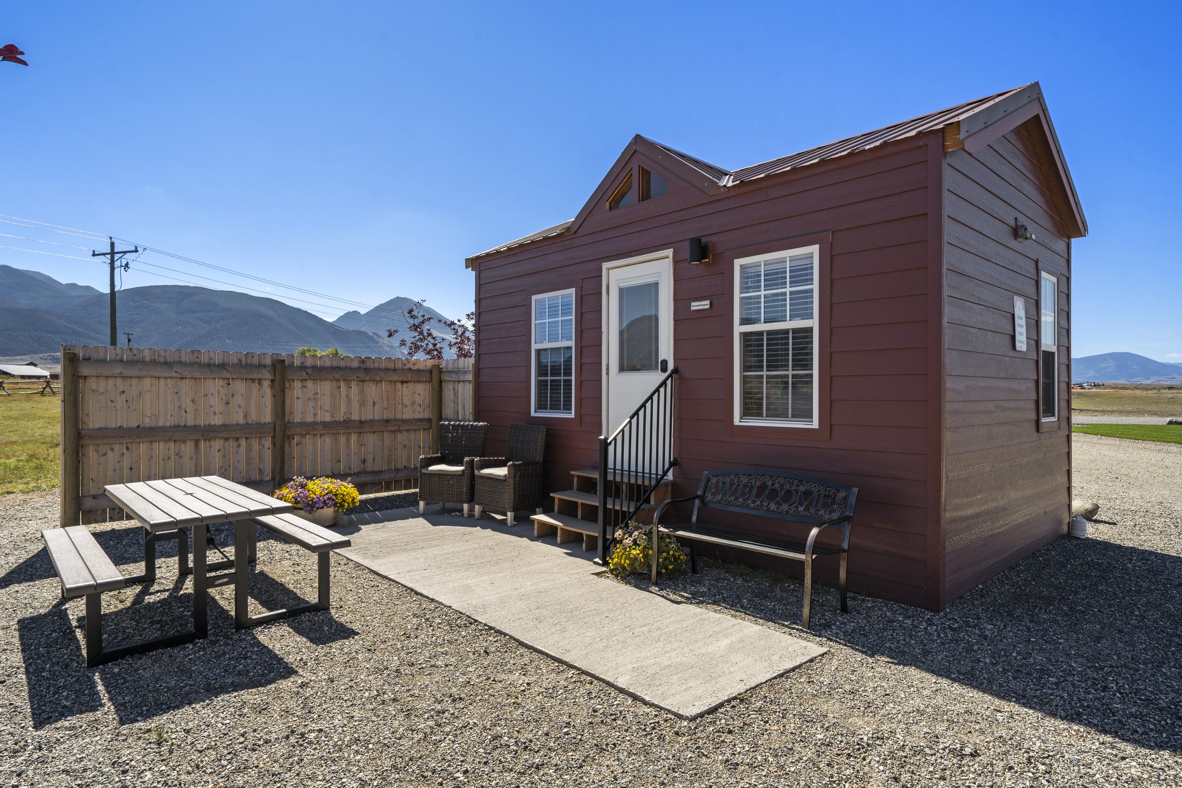 Grasshopper Cabin, with a concrete patio, picnic table, two wicker chairs and a Welcome bench, mountains behind