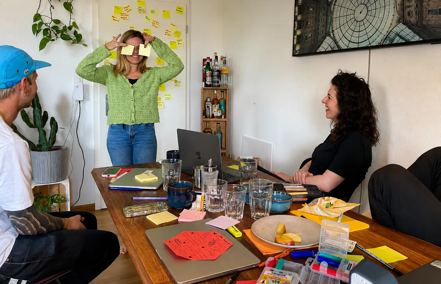 Workshop am grossen Tisch mit Laptops und Post-its, eine Frau steht und hält sich Zettel vor die Augen
