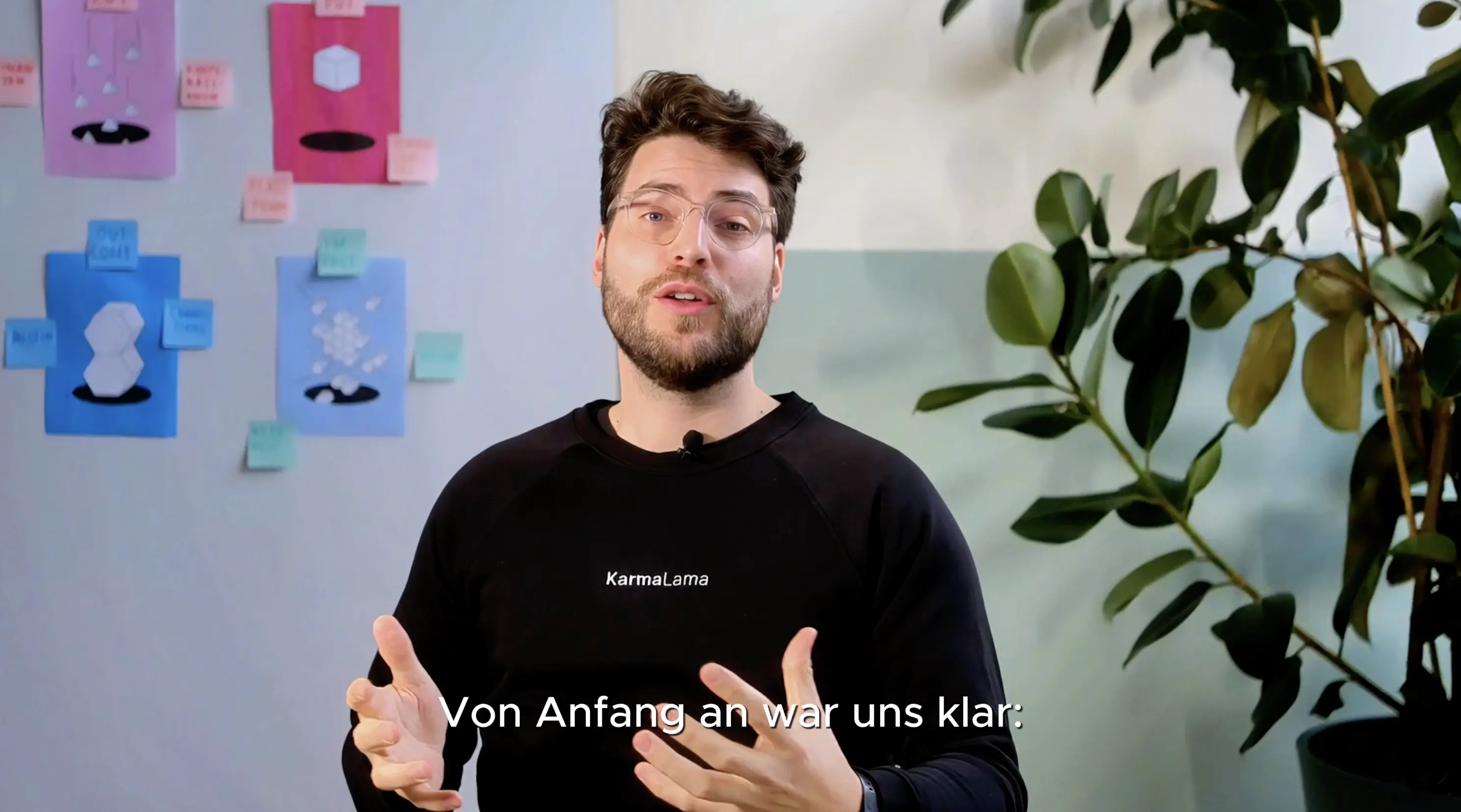 Videostill: ein Mann spricht in die Kamera, im Hintergrund eine Wand mit Illustrationen und Post-its